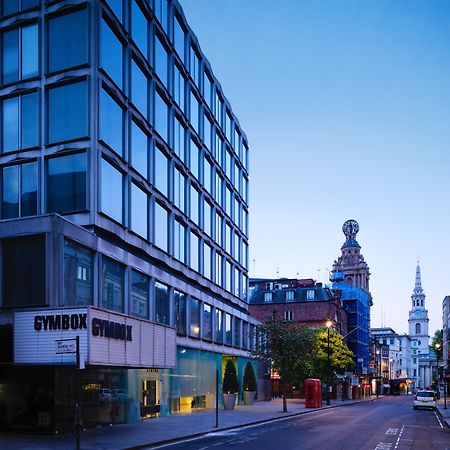 St Martins Lane London Hotel Kültér fotó The photo shows a street scene during twilight, featuring a modern, glass-fronted building on the left side, which appears to be a gym called "GYMBOK GYM." On the right, there are more traditional architectural elements, including a tower and a steep