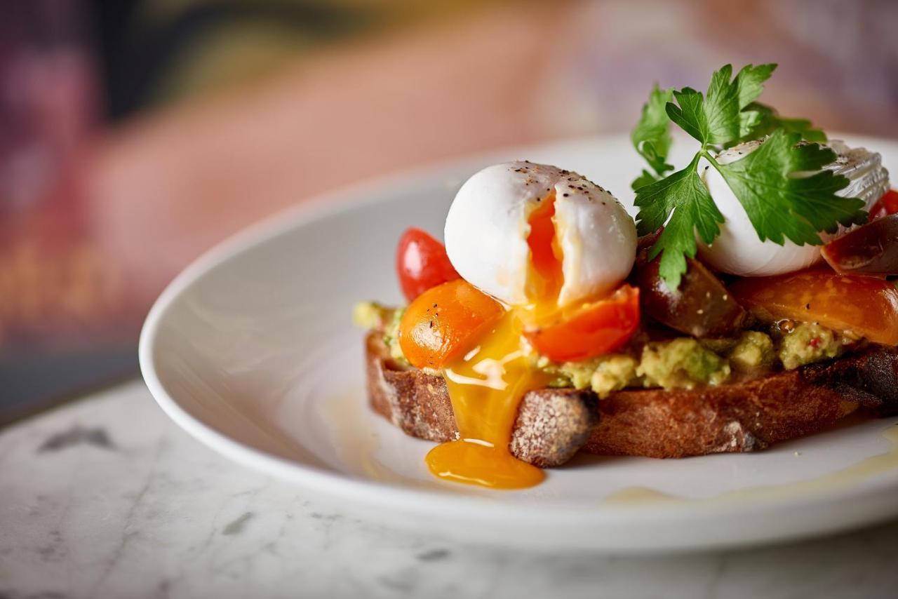 St Martins Lane London Hotel Kültér fotó The photo showcases a beautifully presented dish on a white plate. It features a slice of brown bread topped with a spread that resembles avocado, accompanied by vibrant cherry tomatoes. Atop the bread, there are two poached eggs, with the yolks runn