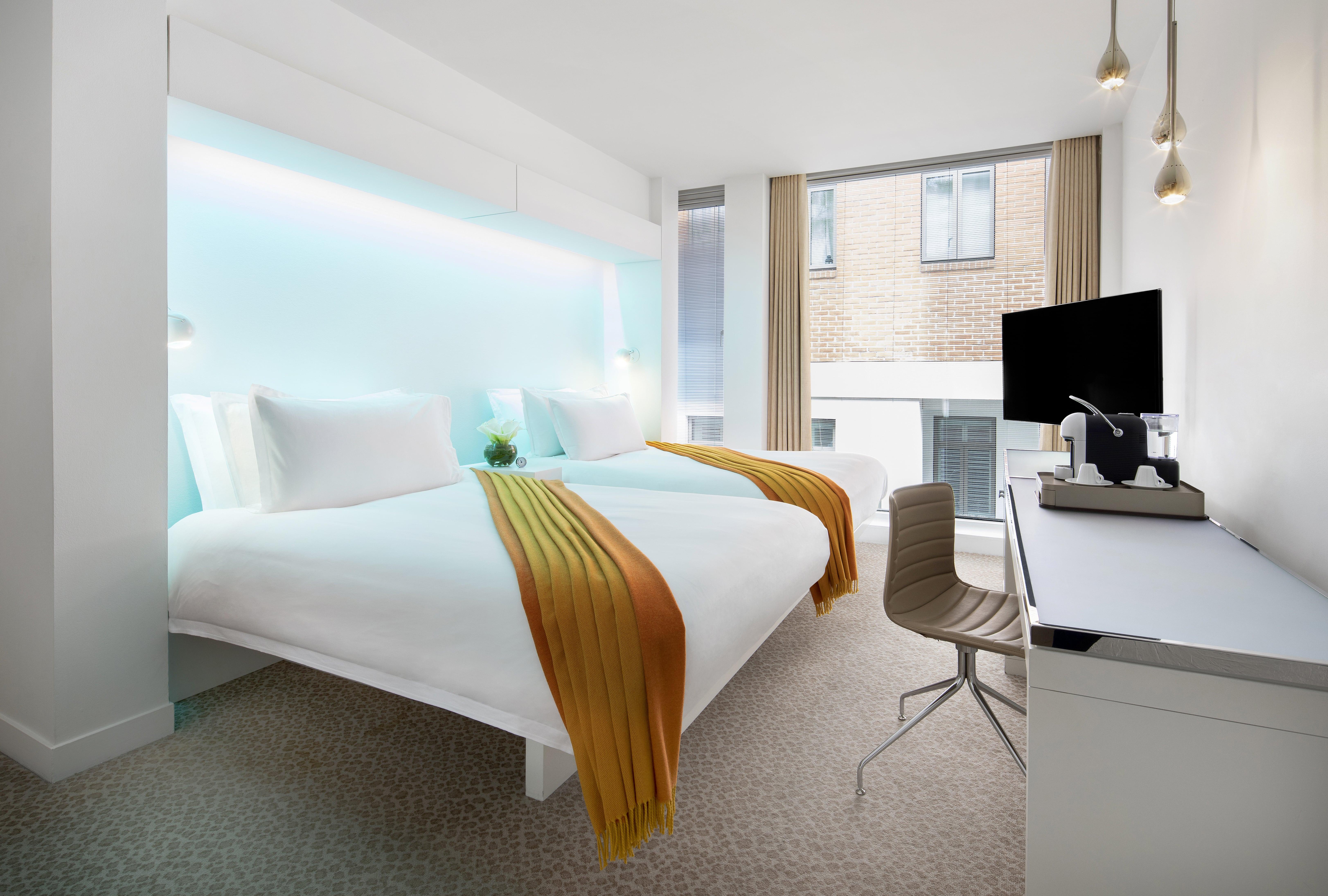 St Martins Lane London Hotel Kültér fotó The photo shows a modern hotel room with a clean, contemporary design. There are two beds, each with white bedding and vibrant orange and yellow throws. Behind the beds, there is a glowing blue accent wall, adding a stylish touch. The room features a