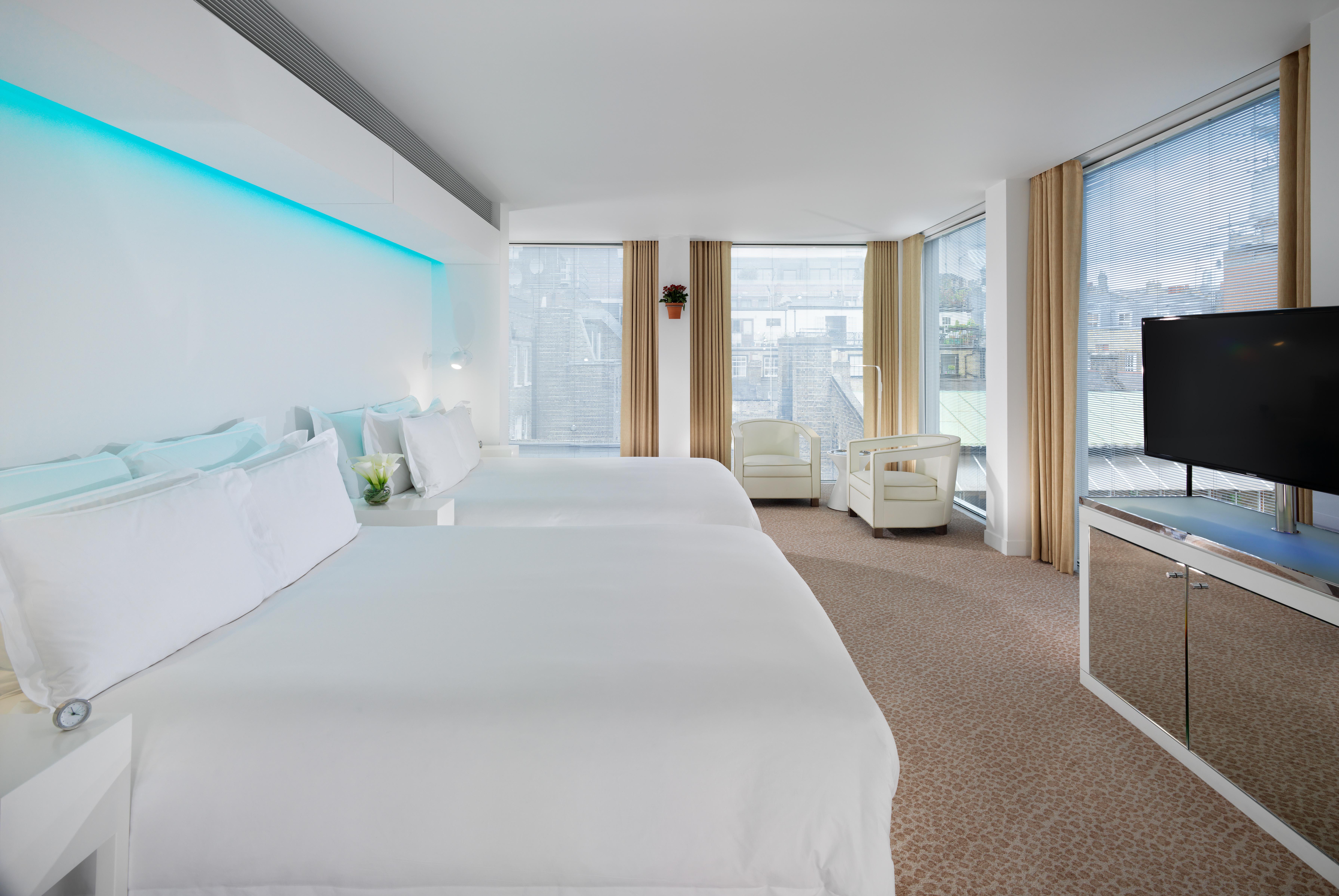 St Martins Lane London Hotel Kültér fotó The photo shows a modern hotel room featuring two large beds with white bedding. The room has large windows with light-colored curtains, allowing natural light to fill the space. There is a small seating area with a white sofa and a bedside table. A 