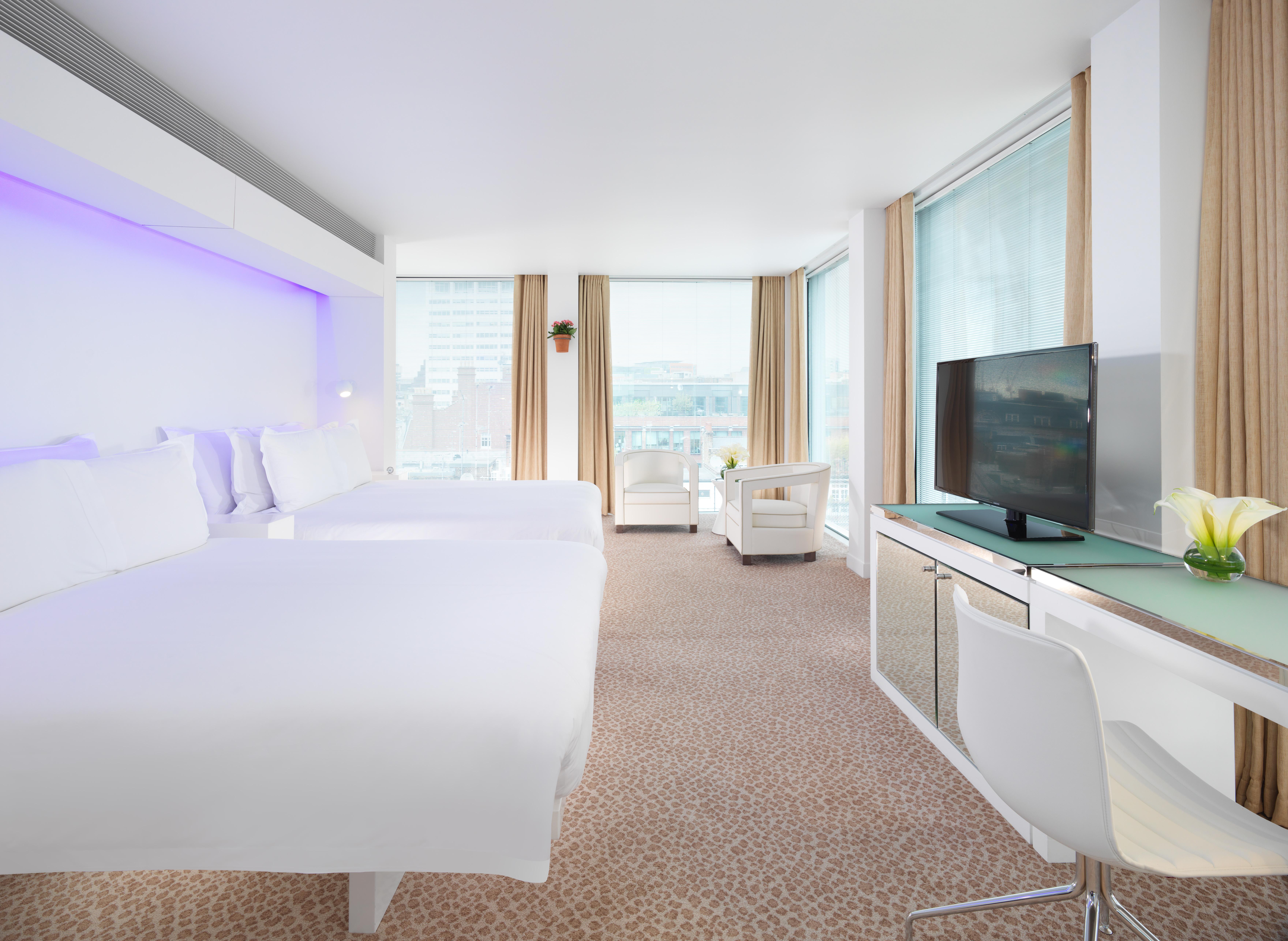 St Martins Lane London Hotel Kültér fotó The photo shows a modern hotel room featuring two beds with white linens, a sleek design, and large windows allowing natural light to enter. The room includes a comfortable chair, a desk with a television mounted on the wall, and a stylish decorative
