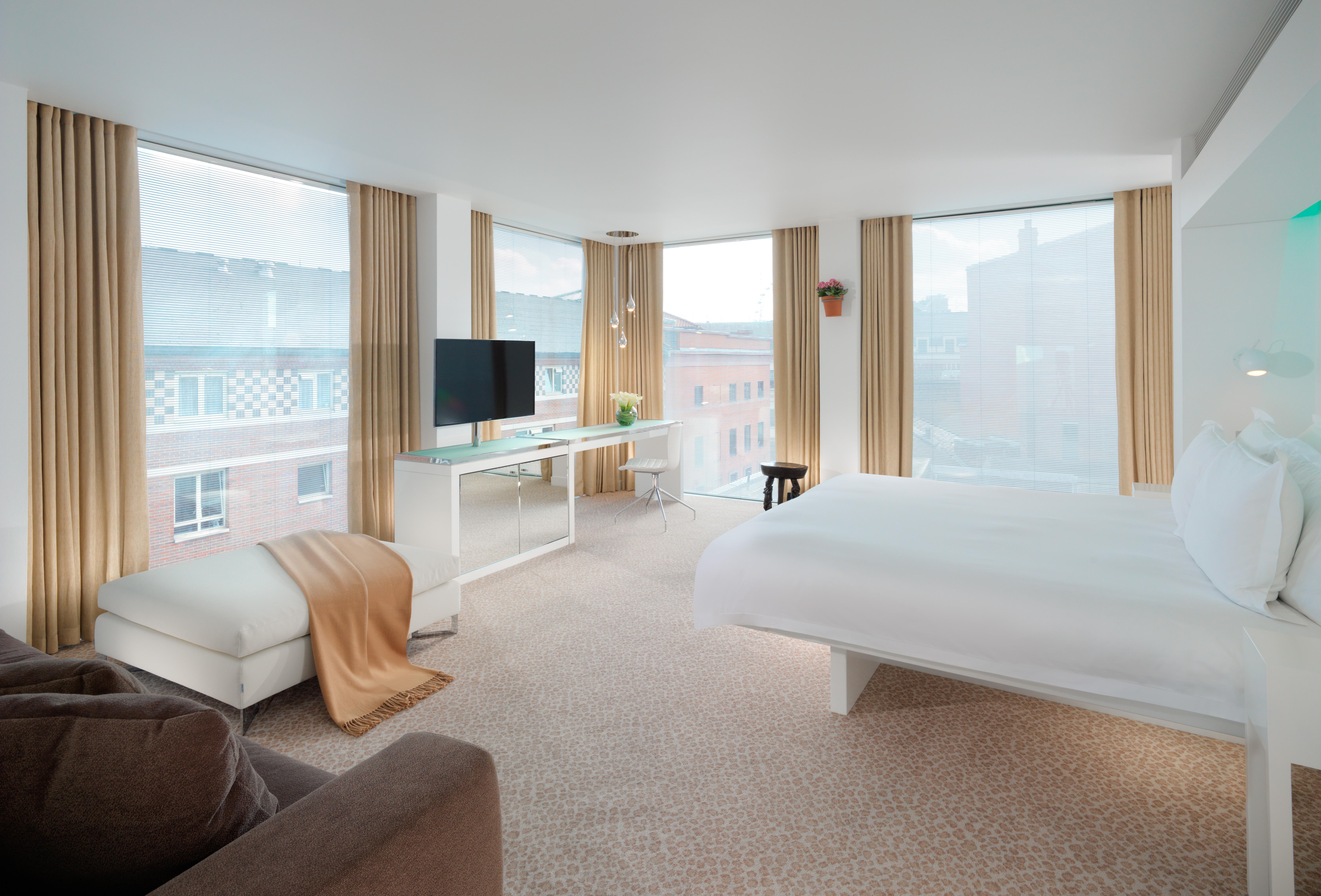 St Martins Lane London Hotel Kültér fotó The photo displays a modern hotel room that features large windows allowing natural light to fill the space. The room includes a comfortable bed with white linens, a cozy chair and a throw blanket in neutral tones, and a small side table. There is a 