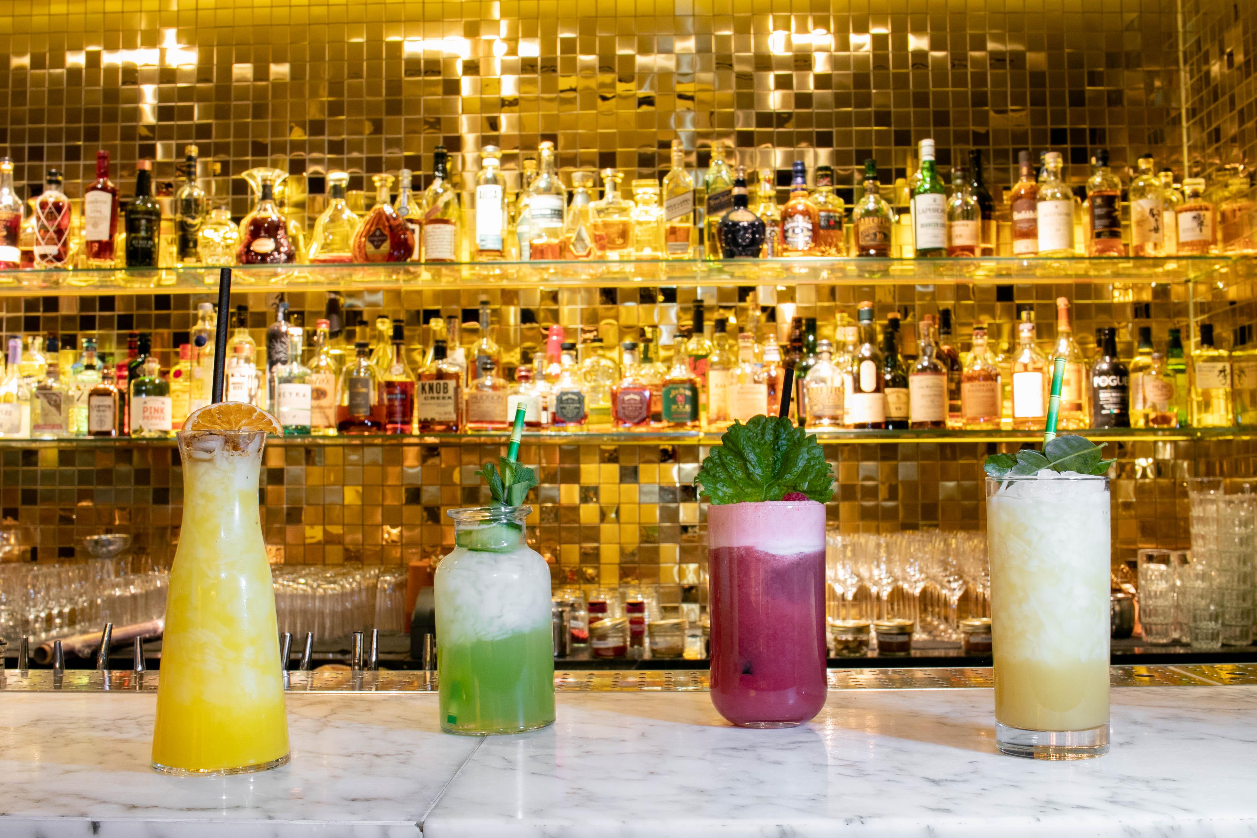 St Martins Lane London Hotel Kültér fotó The photo features a stylish bar scene with four colorful cocktails displayed on a marble countertop. Behind the drinks, there is a sparkling, gold mosaic wall lined with various liquor bottles, creating an upscale ambiance. The cocktails include vib