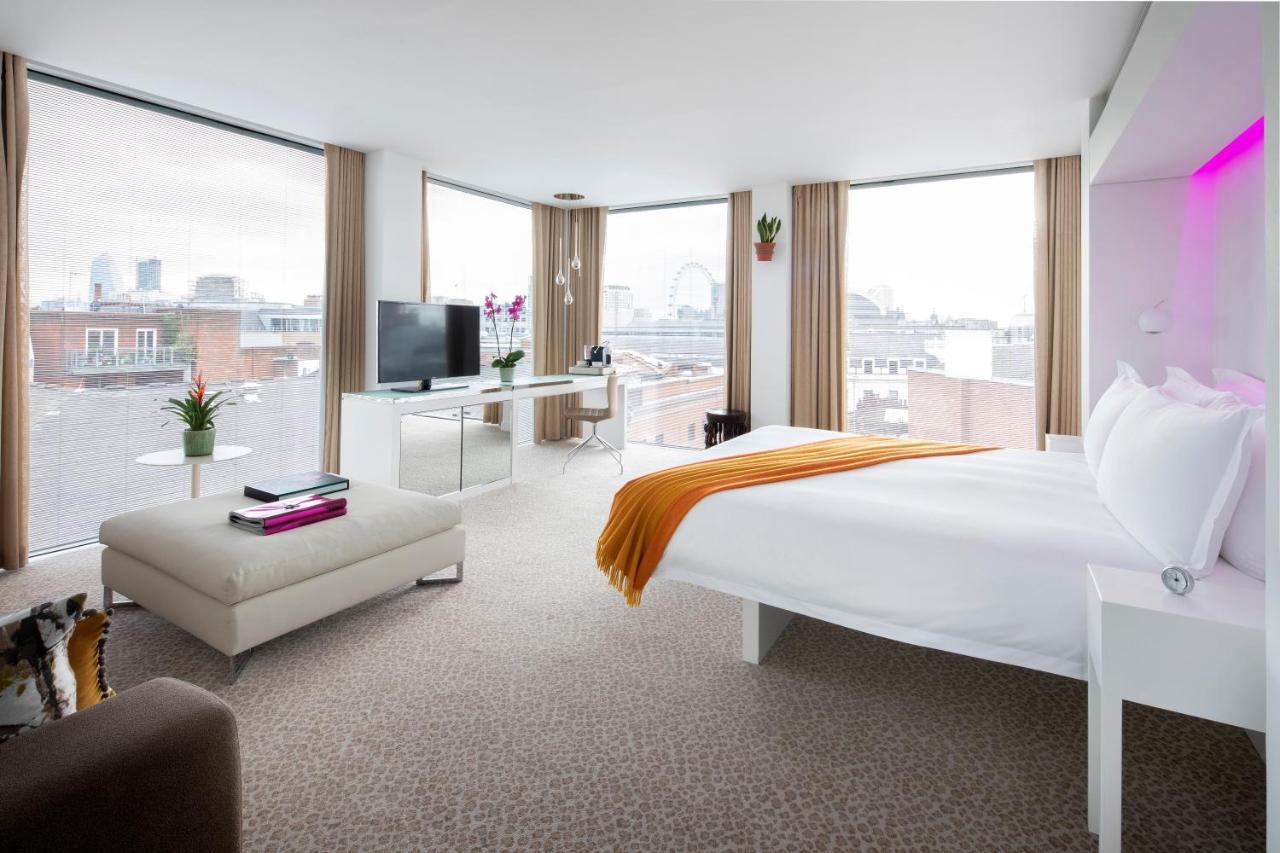 St Martins Lane London Hotel Kültér fotó The photo shows a modern hotel room featuring a large bed with white linens and an orange throw blanket. The room has ample natural light coming through large windows with beige curtains, offering a view of a cityscape. There is a small seating area 