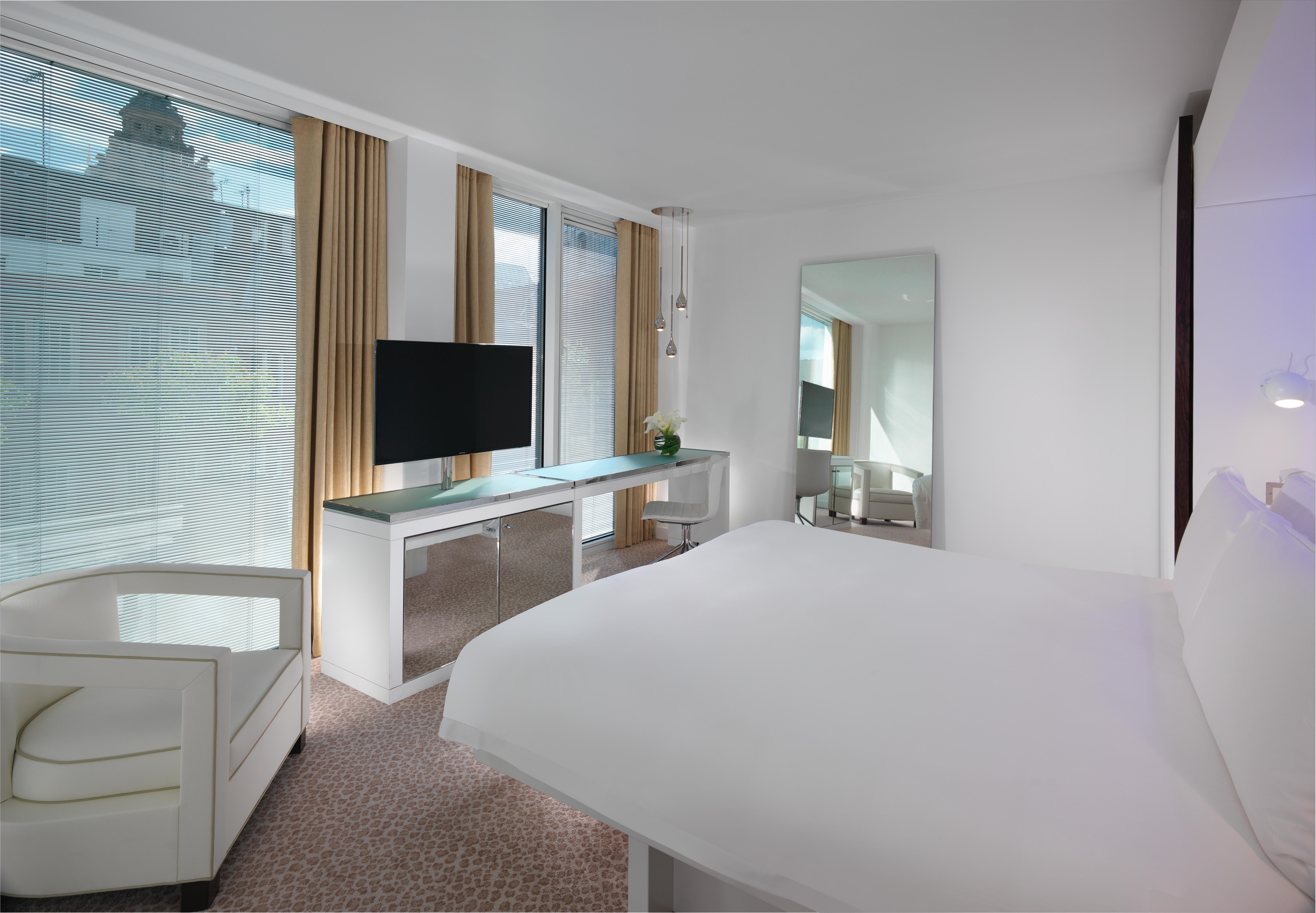 St Martins Lane London Hotel Kültér fotó The photo shows a modern hotel room featuring a king-sized bed with white bedding. There is a sleek, minimalist design with light-colored walls and a carpeted floor. A large window allows natural light to fill the room, with sheer curtains providing 