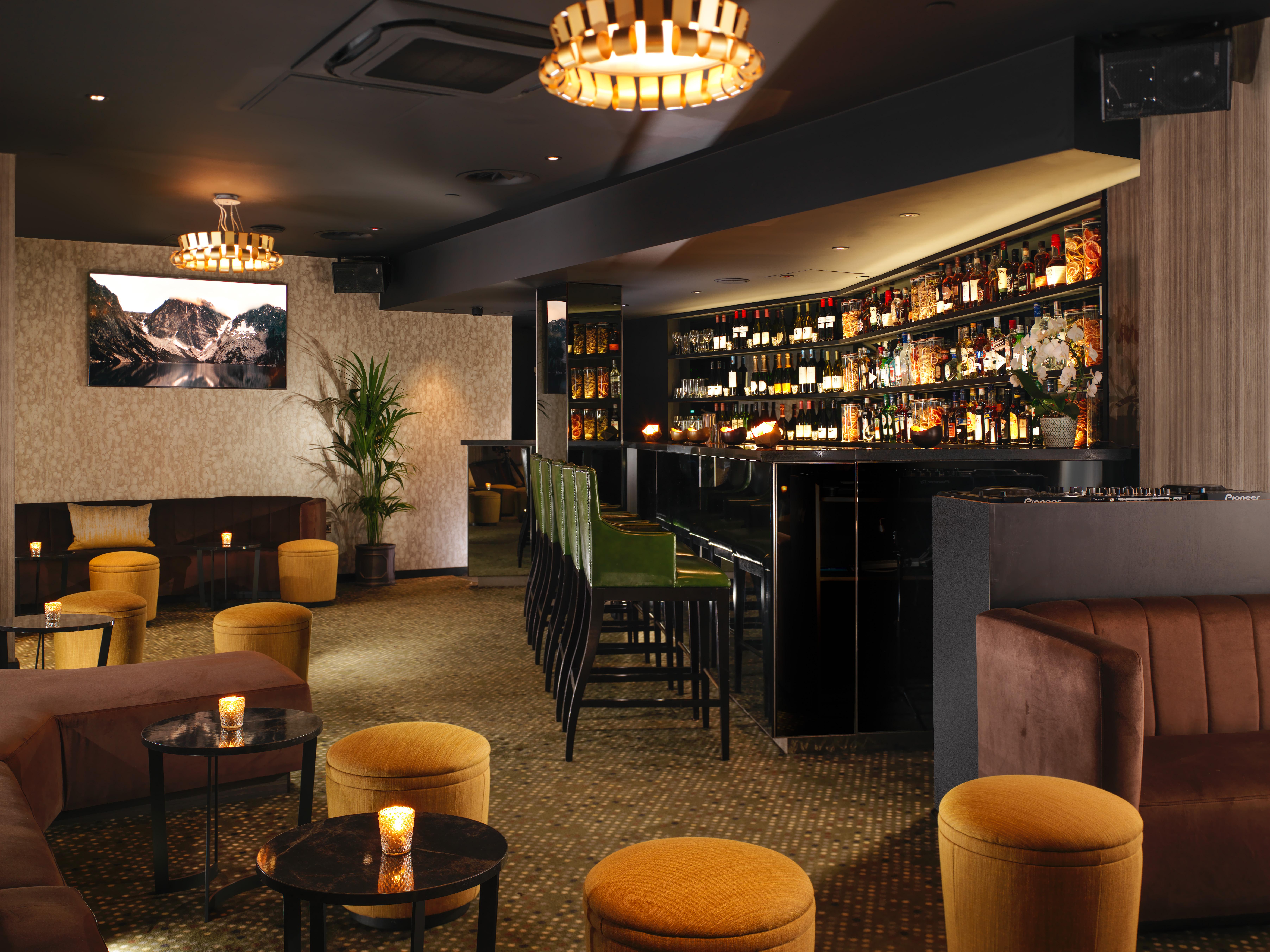 St Martins Lane London Hotel Kültér fotó The photo depicts a stylish bar or lounge interior. The space features a dark, sophisticated color scheme with plush seating, including brown couches and vibrant yellow stools. A illuminated bar area is prominently displayed, lined with various bottl