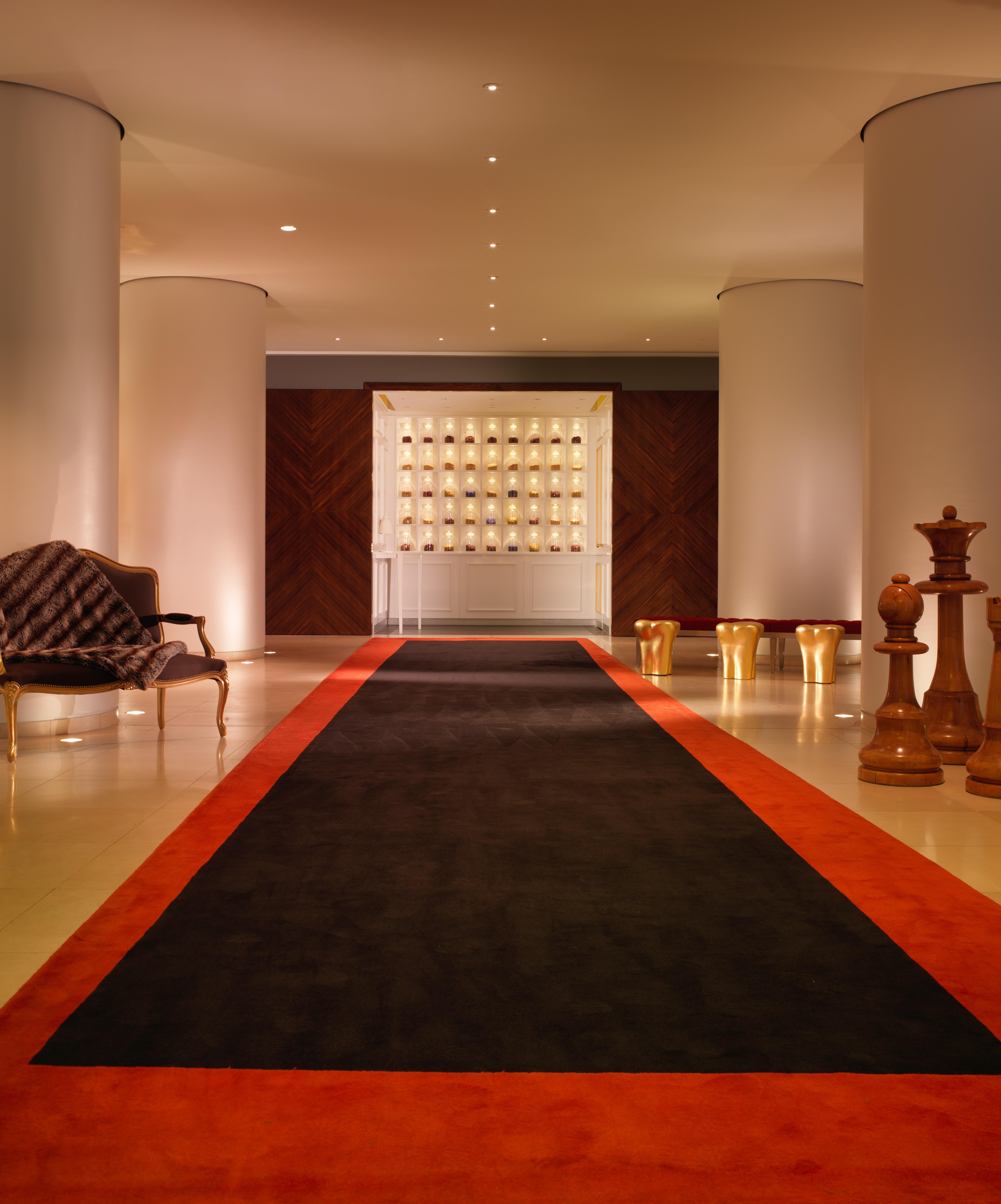St Martins Lane London Hotel Kültér fotó The photo shows a modern, elegant hallway or lobby area. There is a dark carpet runner in the center, bordered by a vibrant orange strip. On the left side, there is a stylish armchair, and on the right, large wooden pieces that resemble chess pieces.