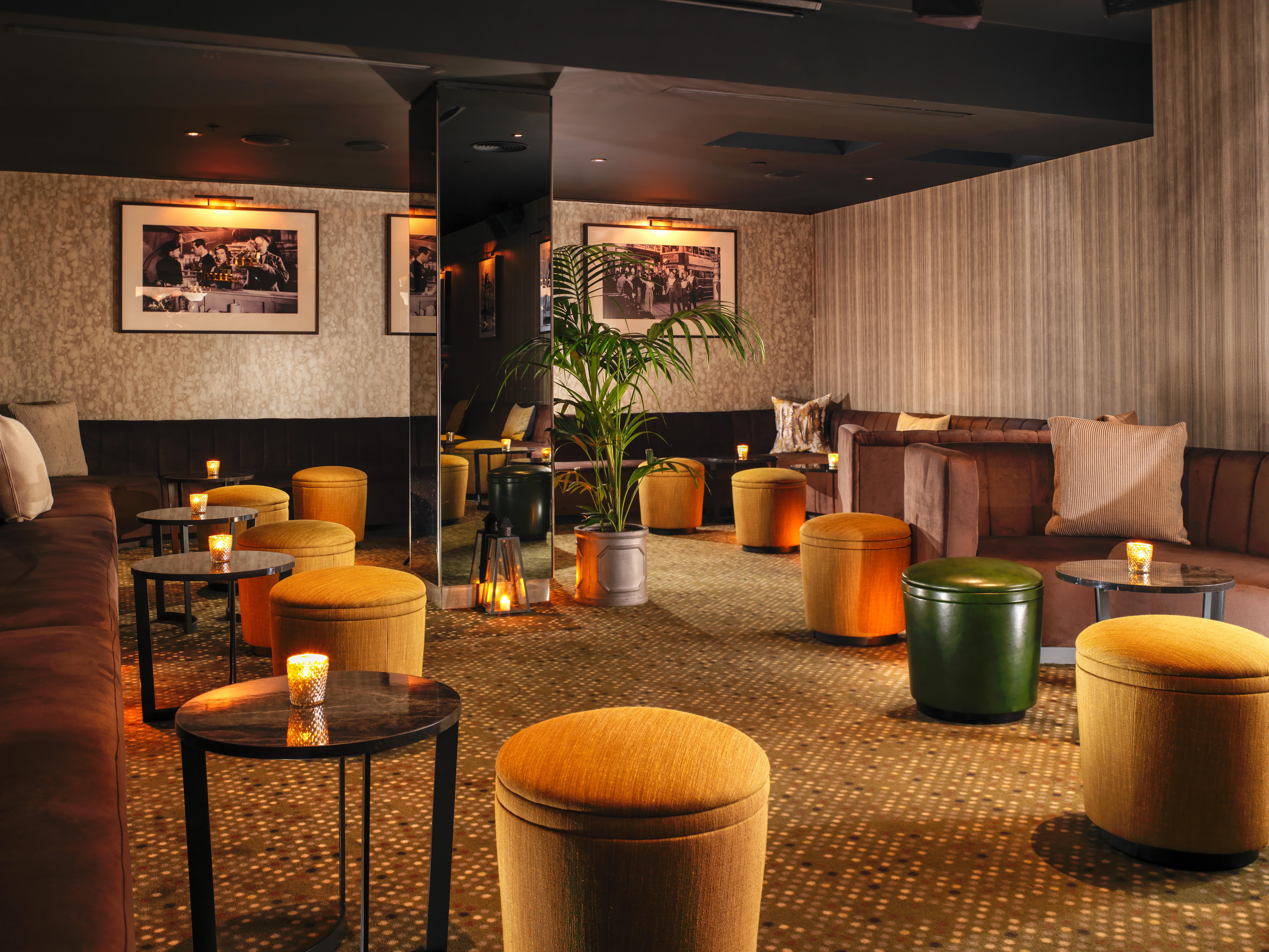St Martins Lane London Hotel Kültér fotó The photo shows a cozy lounge area with a warm and inviting atmosphere. The space features a mix of seating arrangements, including cushioned benches and small round tables. There are several colorful ottomans, predominantly in shades of yellow and g