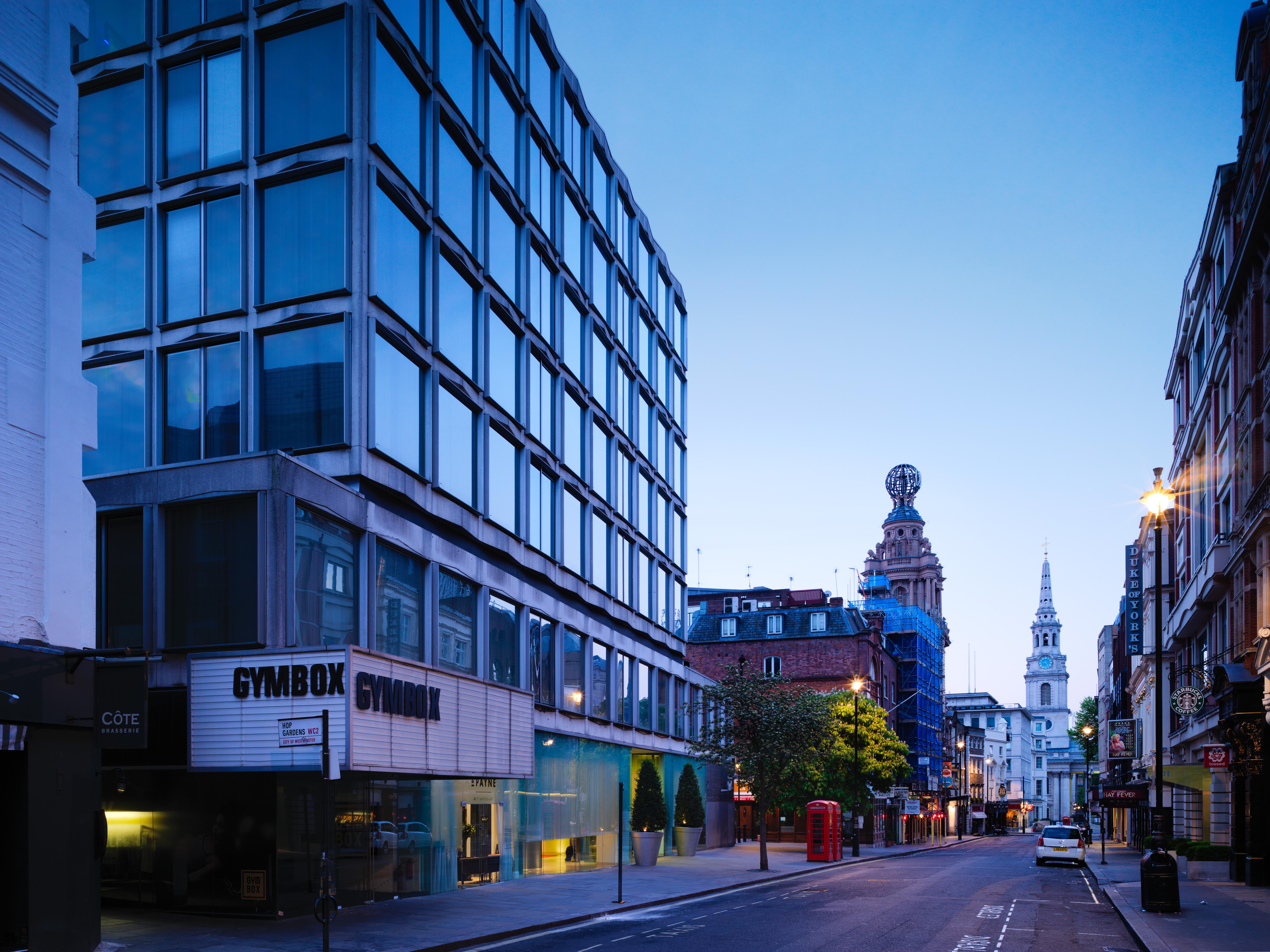 St Martins Lane London Hotel Kültér fotó The photo shows a street scene during twilight, featuring a modern, glass-fronted building on the left side, which appears to be a gym called "GYMBOK GYM." On the right, there are more traditional architectural elements, including a tower and a steep
