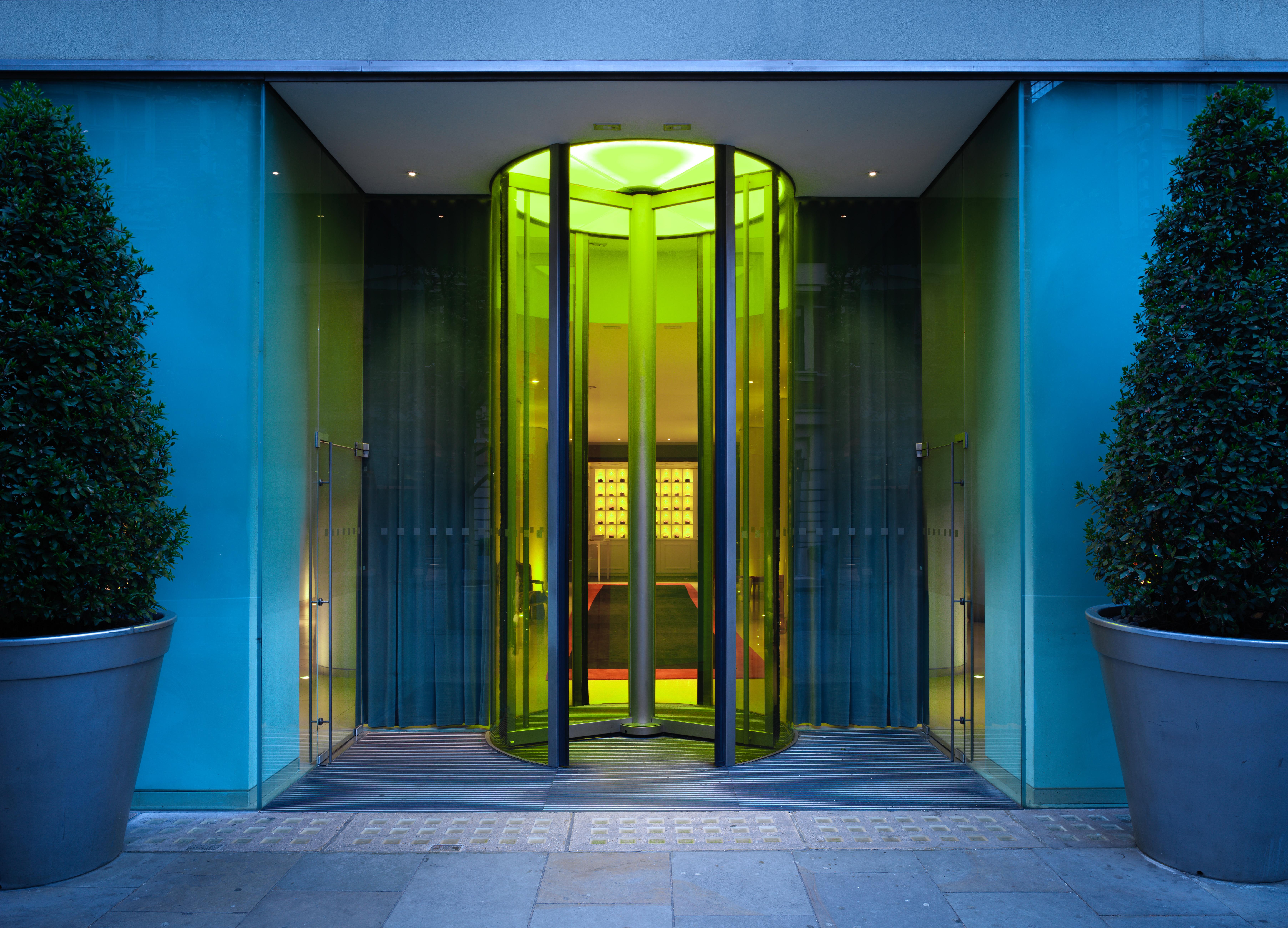 St Martins Lane London Hotel Kültér fotó The photo shows a modern glass entrance with a revolving door. The door is surrounded by sleek glass panels that reflect light, creating a vibrant ambiance. The entrance is complemented by large, symmetrical planters containing rounded shrubs on eith