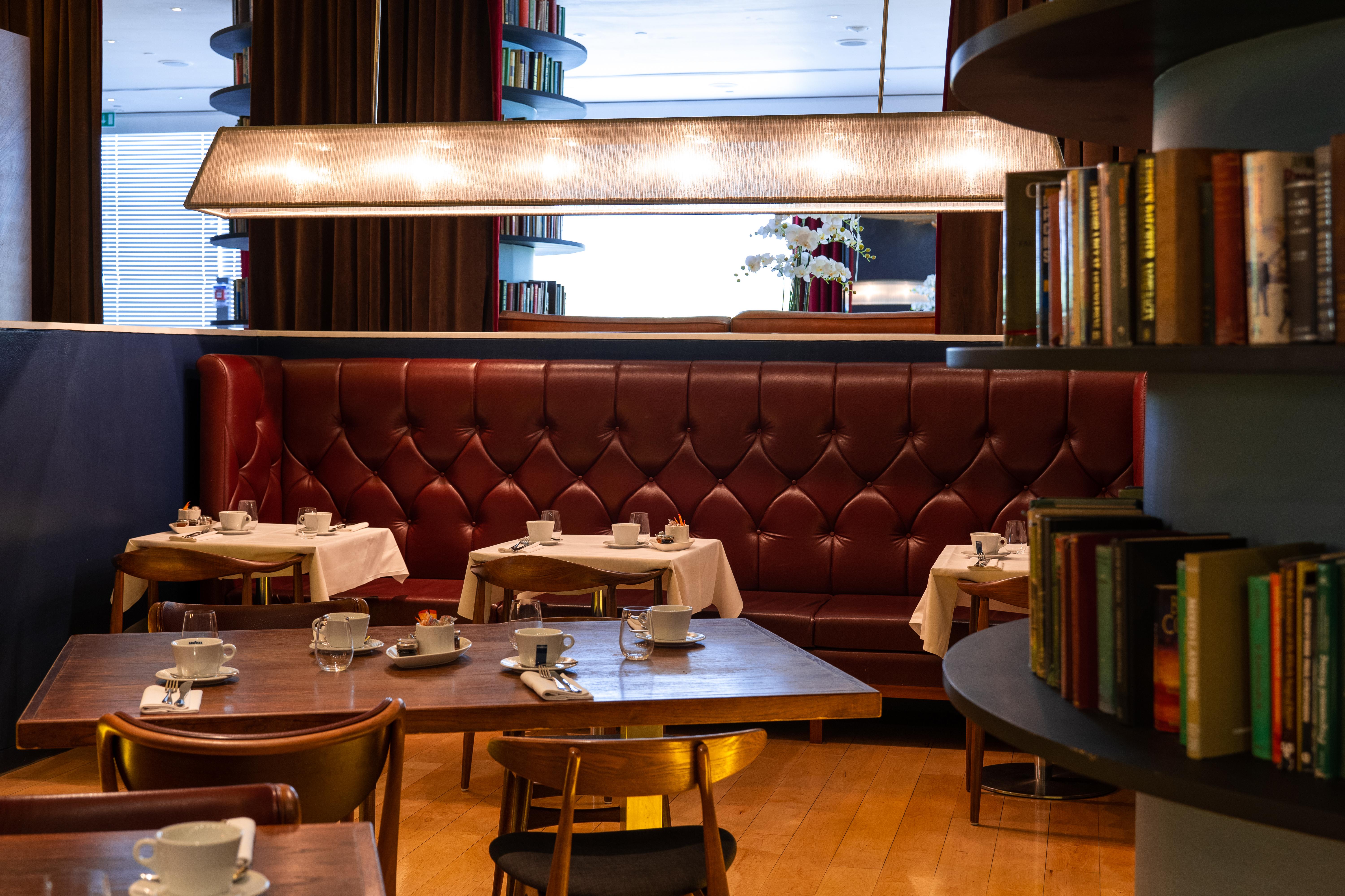 St Martins Lane London Hotel Kültér fotó The photo depicts a cozy, elegantly designed restaurant interior. There are several round tables set with white tablecloths and accompanied by small white cups, likely for tea or coffee. The seating includes a plush, burgundy leather banquette agains