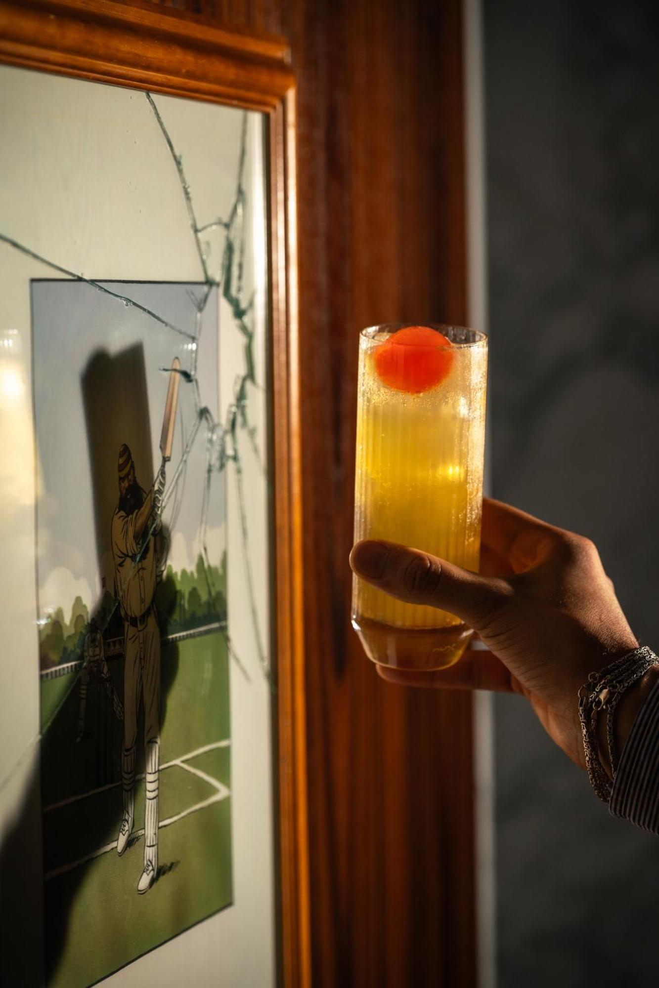 St Martins Lane London Hotel Kültér fotó The photo shows a hand holding a tall glass containing a drink, which appears to be a yellow cocktail topped with a bright red cherry. In the background, there is a piece of art or a photograph that features a figure, possibly in a baseball or sporti