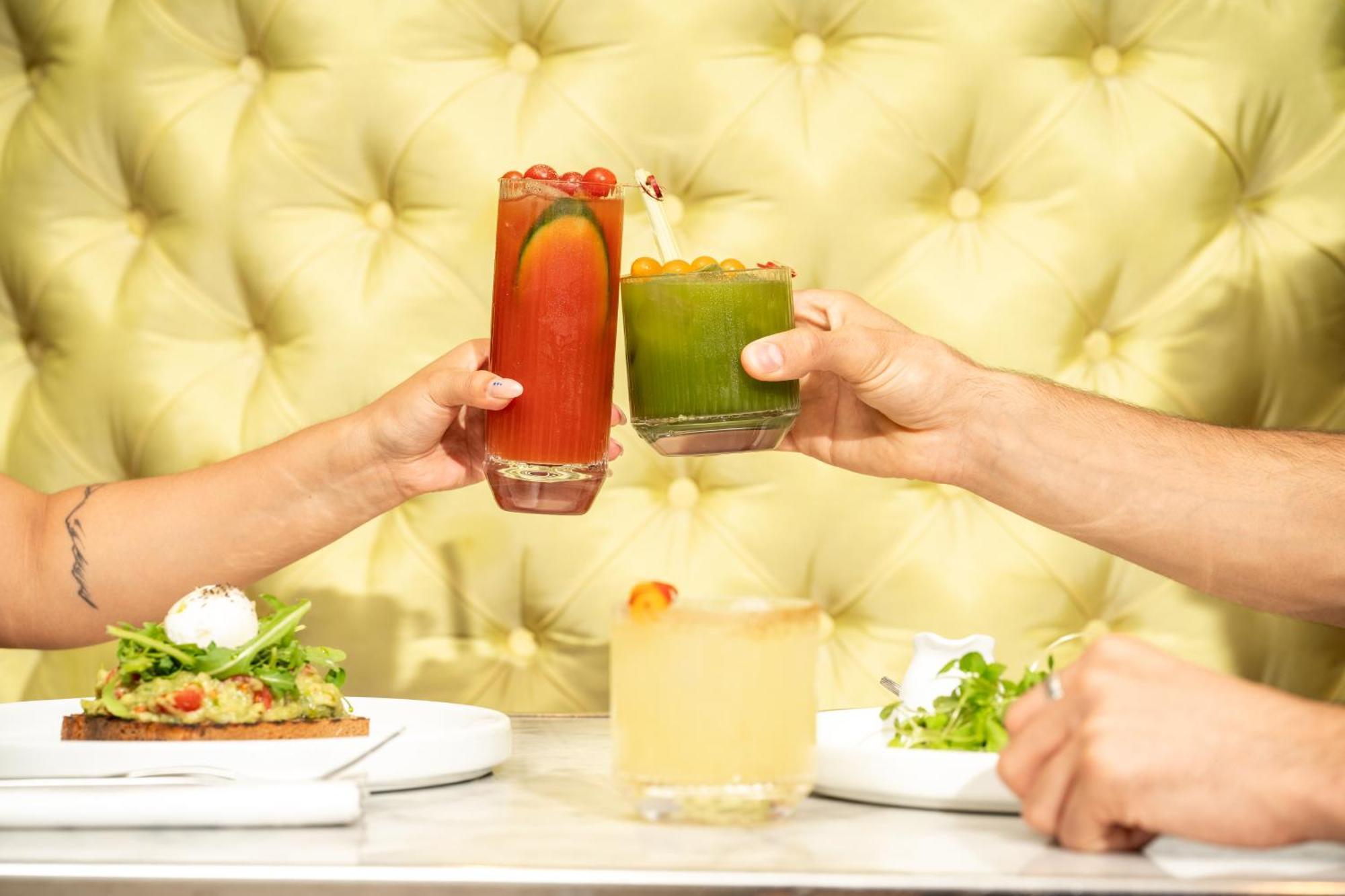 St Martins Lane London Hotel Kültér fotó The photo features a vibrant dining scene with two hands clinking glasses in a toast. One glass contains a red drink, possibly a cocktail, adorned with garnishes, while the other is a green beverage, likely a smoothie or juice. In the foreground, the