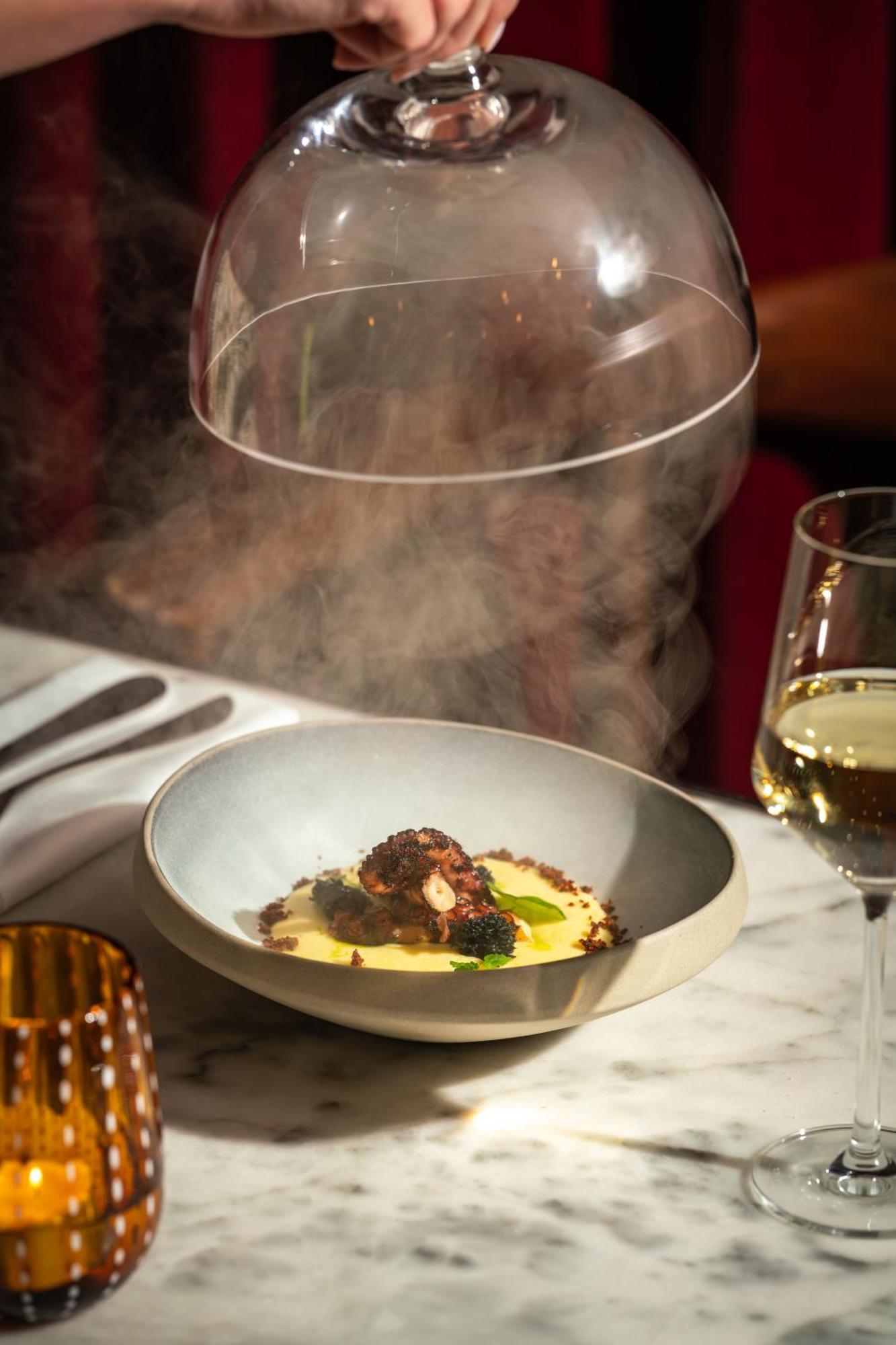 St Martins Lane London Hotel Kültér fotó The photo depicts an elegant dish served in a shallow bowl. The main feature appears to be octopus, garnished with greens, and placed on a smooth, creamy base, possibly a type of puree. A dome is being lifted from above the dish, releasing steam, whi