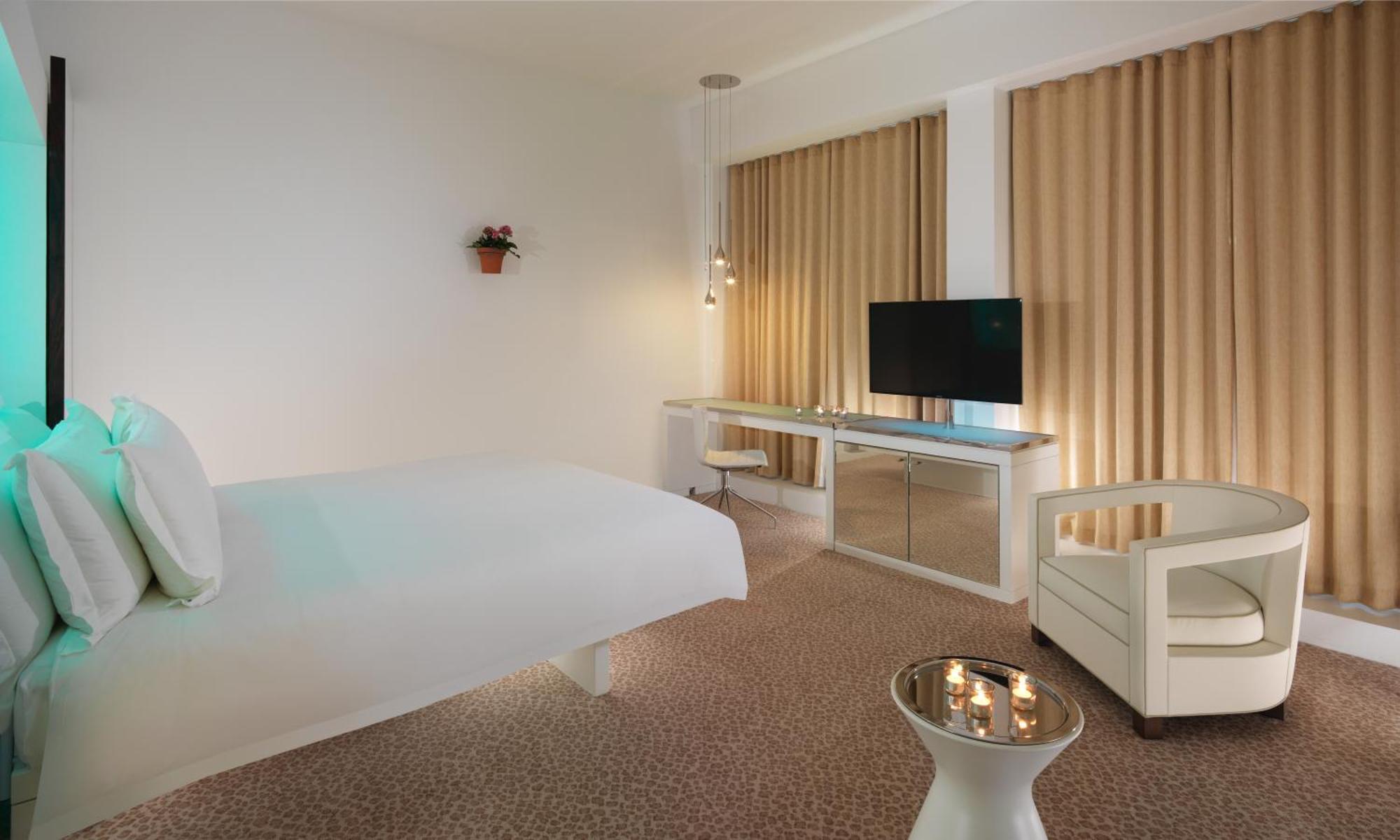St Martins Lane London Hotel Kültér fotó The photo shows a modern hotel room with a minimalist design. It features a large bed against one wall, a flat-screen TV mounted on another wall, and a contemporary desk with a mirror. Large windows are dressed with light-colored curtains, allowing n