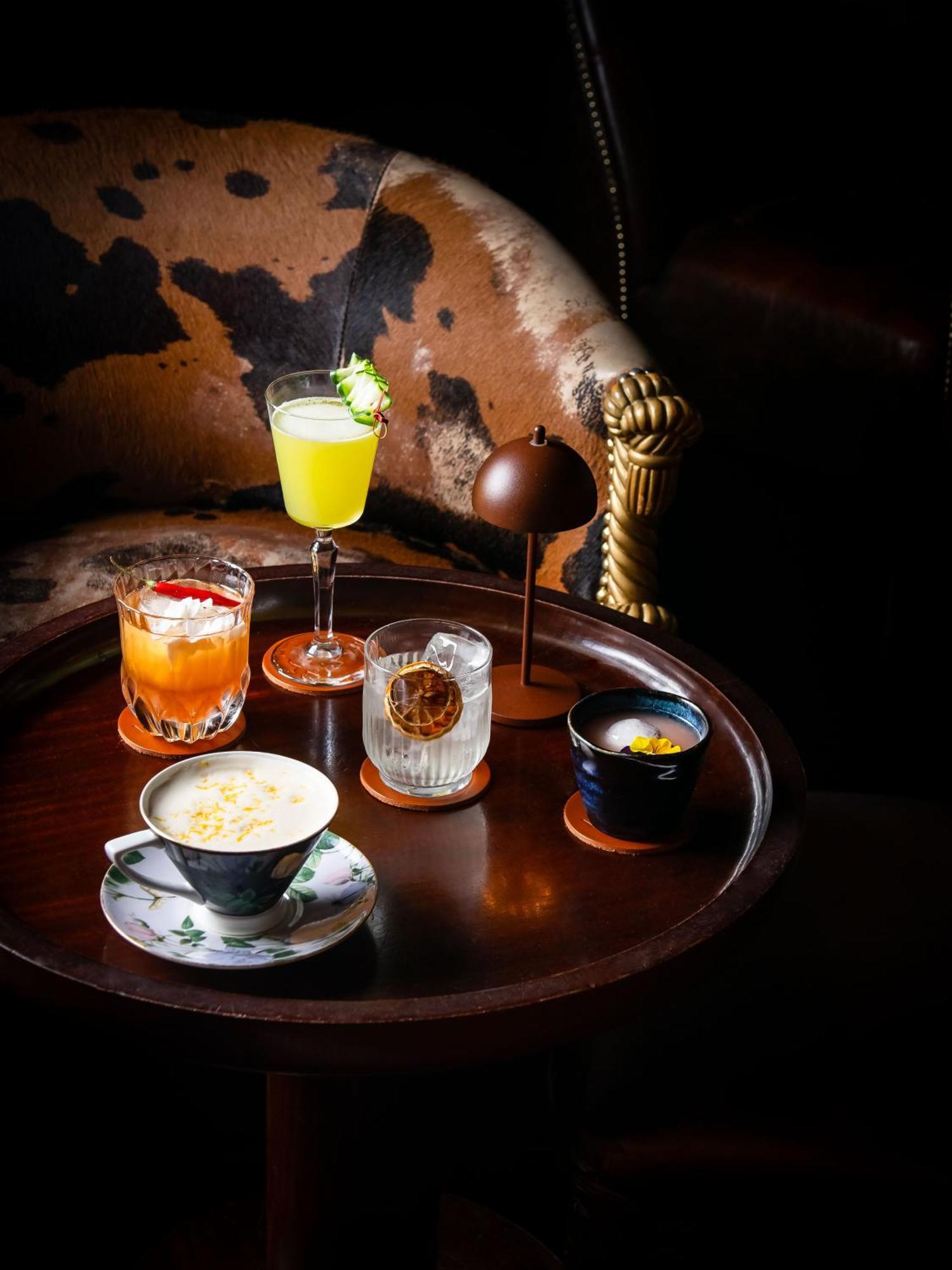 St Martins Lane London Hotel Kültér fotó The photo displays a variety of drinks arranged on a round wooden tray. There are five distinct beverages:

1. **A light green cocktail** in a tall glass, garnished with a slice of fruit, suggesting a refreshing flavor.
2. **An orange drink** served 