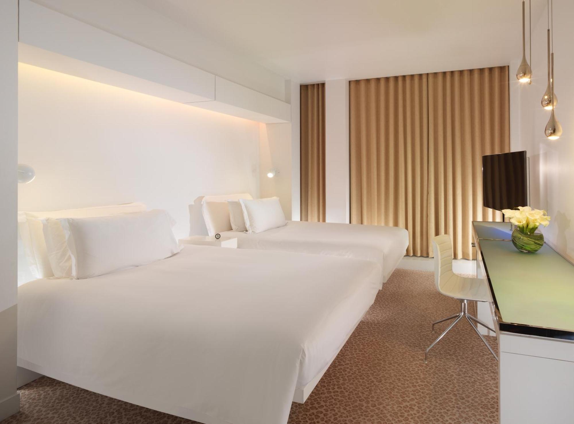 St Martins Lane London Hotel Kültér fotó The photo shows a modern hotel room featuring two white beds neatly made with minimalistic design. The walls are primarily white with warm lighting highlighting the space. There are large windows dressed with light brown curtains. A small desk with a
