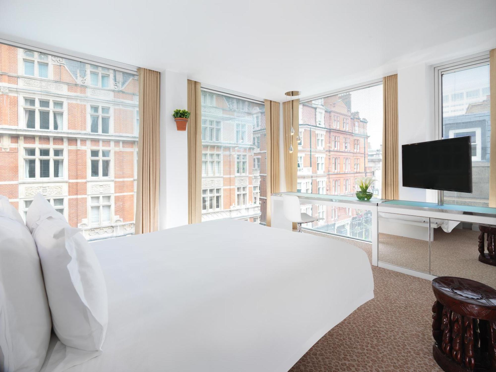 St Martins Lane London Hotel Kültér fotó The photo shows a modern hotel room featuring a large bed with white linens, positioned near large windows that provide abundant natural light. The windows offer a view of brick buildings outside. There is a flat-screen TV mounted on the wall, a mini