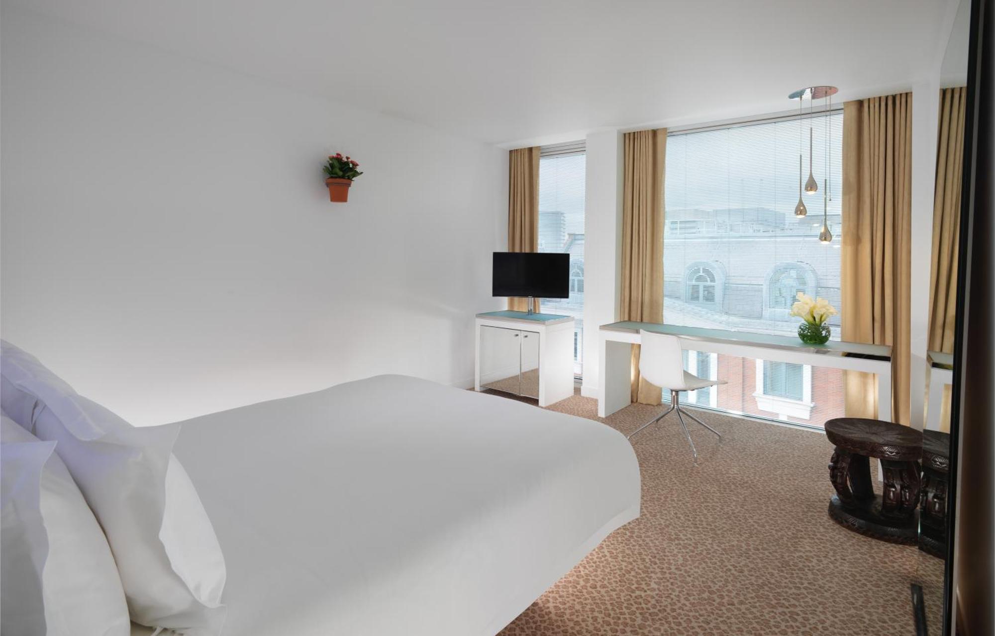 St Martins Lane London Hotel Kültér fotó The photo shows a modern hotel room that features a large bed with white linens, a small desk with a chair, and a television mounted on the wall. There are large windows covered with light curtains, allowing natural light to fill the room. A small po